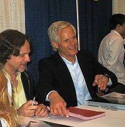 The image is of two men sitting at a table signing items. The one on the left is wearing a golden shirt and has brown hair and a mustache. The one on the right is looking up and has white hair.