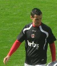 A young man in black and red association football clothing walks across the viewer's line of sight, looking to his left. There is a shadow across his body.
