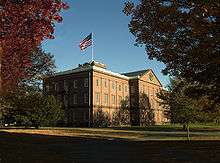 An old brick building in the Georgian architectural style surrounded by a wide lawn and trees. An American flag flies over the center of the building.