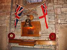 The White Ensign and Union Flag are crossed together on an interior stone wall above a brass bell and wooden plaque. Beneath this is a glass and wood cabinet. Poppy wreaths lie to either side.