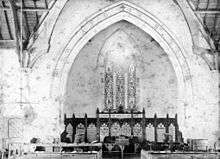 Black and white photograph, originally on sensitized albumen, depicting the eastern end of the church interior. The lettering around the arch begins "O Come Let Us Worship And Fall Down", but the photographic materials used make the blue letters difficult to make out.