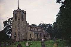 A small church with a square Ashlar tower, in the Early English style. The tower is to the left, the nave to the right, and we are looking somewhat obliquely at it.  The church is ringed by medium size trees, and a large Yew dominates the right of the picture, in the middle distance.  A handful of mismatched gravestones dot the grassy nearground of the picture.