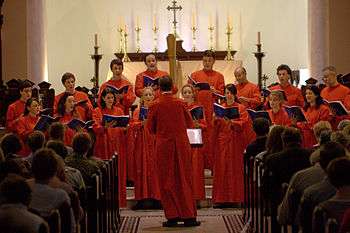 A photo of a red-robed choir in the chancel with the altar behind them. Candles and a brass cross are on the altar and the choirmaster is facing the singers with his back to the camera