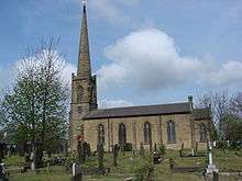 Under a partly cloudy sky is a traditional English church pictured from the side. On the right are a large, square tower and a tall spire with a weather vane atop. In front of the church is a graveyard with an assortment of gravestones and monuments, and some trees with early spring foliage.