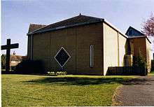 A modern Church of England church. A large concrete crucifix, of similar height to the church, is beside the building.