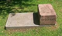 A block of orange-pink stone on a concrete bed, surrounded by grass, with the name "Stallard" carved into the side facing the viewer