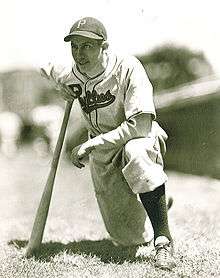 A baseball player is kneeling with one knee while leaning onto a baseball bat.