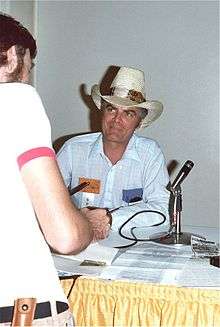 Man in a cowboy hat and white shirt with thin blue stripes sitting at a table with a microphone