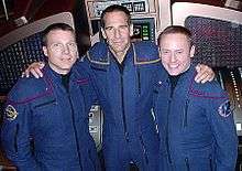 Three men in blue jumpsuits are arm in arm.