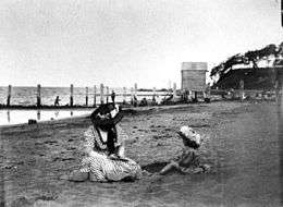 Woman and young child sitting on the beach at Sandgate. The woman appears to be reading to the child, and both are wearing hats. A boatshed and jetty are visible in the background. People are fishing from the jetty.