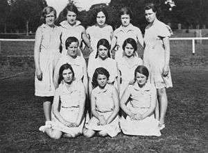 A black-and-white photo of a netball team. All the girls in the picture are school aged, wearing white and have their netball skirts on.