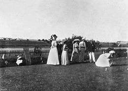Men and women play lawn bowls. The women, one of whom is about to bowl a ball down the green, wear large hats and long dresses. A scoreboard has been set up next to the group.