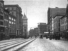 A black and white photograph showing the post office tower in the middle of other buildings on one side of a street with tracks down the middle