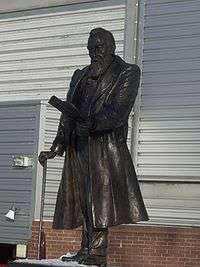 A bronze statue of a man with a large beard, wearing a long coat, leaning on a walking stick, and holding some papers