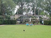 a grassy lawn in front of a statues complex several people sunbathe or sit in the midground facing in the background a group of white statues that are significantly larger than the people. In the foreground a black bird is walking away from the whole tableaux