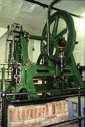 Green painted machinery with a wheel at the top anmd a small brick wall in front
