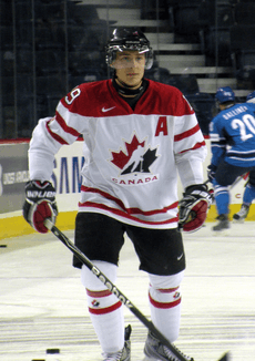 Hockey player in white Canada uniform. He stands on the ice, hands by his side, one holding his stick.