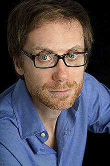A blue-eyed, ginger-bearded man in a blue shirt and glasses looking into the camera before a black background.