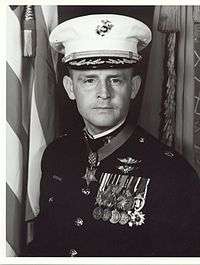 A black and white image of Pless wearing his military dress uniform with hat and medals. His Medal of Honor is visible around his neck and there is an American flag in the background.