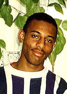 Photograph of a young Afro-Caribbean male, cropped to show his chest and head. He has black hair, shaved very short, and a slight moustache. He is wearing a navy-and-white vertically striped crew neck shirt. He is standing in front of a large indoor plant.