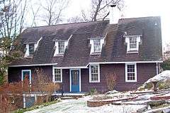 A brown house with low sloping black roof and dormer windows. There is a light snow covering on the brick and stone fountain and front lawn.