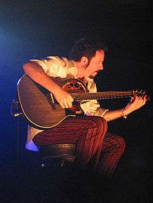 Lukather sits on stage, illuminated by a spotlight, playing a dark grey Ovation Adamas acoustic-electric guitar.