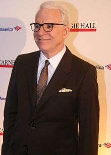 A man in his middle sixties is standing up against a wall. He wears a suit and glasses.
