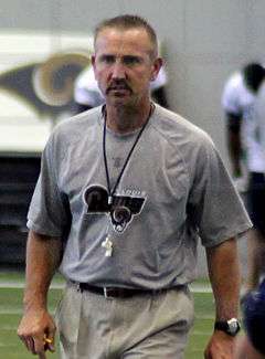 Candid waist-up photography of Spagnuolo on the field in the St. Louis Rams indoor practice facility wearing a grey Rams t-shirt and khaki pants with a whistle hanging from a string around his neck