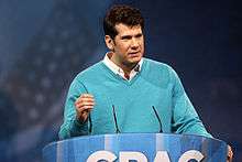 A photo of Steven Crowder at the 2013 Conservative Political Action Conference, wearing a blue sweater over a white collared shirt, speaking at a blue CPAC podium.
