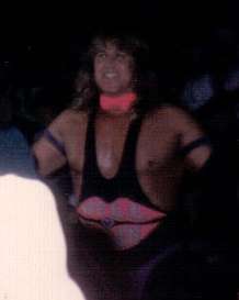 A man with long, wavy brown hair is smiling. He is wearing black wrestling tights with straps that go up over each shoulder. The front of the tights has a large picture of pink lips with the words "Well Dunn" written on them; above the tights, he is wearing purple thong underwear. He also has a pink bow tie around his neck and purple armbands around both biceps.