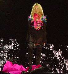 A blond woman standing onstage. Her wavy hair falls on her face. She wears a short black kaftan, a number of pink threads around her neck and long black boots. Near her feet a pink and black cloth is billowing. Behind her, image of water splashing is visible on screen.