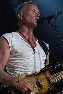 A man in a white shirt standing behind a microphone stand and holding a guitar