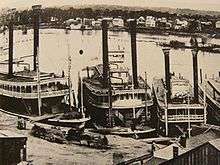 A black-and-white image showing a large river with four steamships docked by the shore. Numerous wooden buildings appear on both sides of the river.