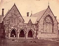 1870s photograph of church-like stone building, with students lying on the grass in front