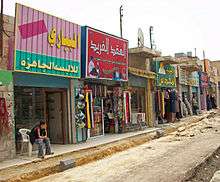 Stores with signs in large Arabic lettering stretching from the lef foreground to the right rear on a street where reconstruction work is in progress