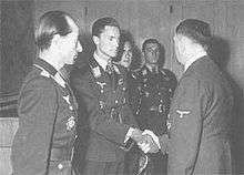 Four men all wearing military uniforms and decorations standing in row. The second man from the left is shaking hands with another man whose back is facing the camera.