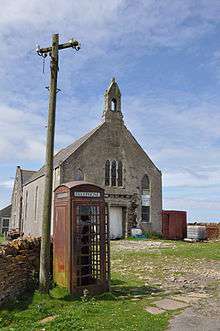 View of Stroma's church, a large stone building with a small tower, with a wrecked red telephone box in the foreground. Next to it is a telephone pole with no wires on it.