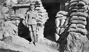 Soldier in peaked cap and puttees outside a sandbagged entrance.
