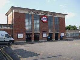 A box-like red-brick building with a projecting flat concrete roof and concrete band below. Four vertical glazed panels in two pairs divide the front elevation. In the centre is the Underground roundel of a red ring with a blue bar and the word "UNDERGROUND". "SUDBURY TOWN STATION" is in lettering fixed to the concrete band beneath the roof.