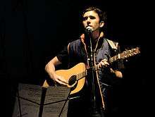 Sufjan Stevens plays guitar in front of a music stand and sings into a microphone