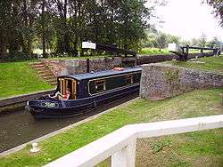 Narrow boat (named Toad) emerging from lock with black gates and white ends of the gate arms. Around the lock is a grassy area.