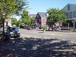 A street scene. Colonial-style buildings frame a wide cobblestone roadway.