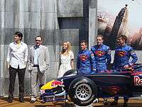 Brandon Routh, Kevin Spacey and Kate Bosworth alongside pilots Christian Klien, Robert Doornbos and David Coulthard, who wear racing overalls with the Superman insignia on the chest. In front of them is the Red Bull Racing racecar, which has the Superman insignia painted atop and sideways of the chassis.