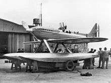 A single-engined monoplane seaplane aircraft is mounted on a wheeled trolley, ten men are standing by the aircraft with one looking into the opened cockpit. The aircraft has 'S1596' painted on the tail.