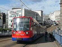 A Sheffield Supertram in current blue, orange and red Stagecoach livery. The tram shown is crossing Park Square bridge and Fitzalan Square and Castle Square can be seen in background, as can tram tracks and numerous commercial buildings.