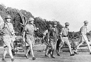 Four British soldiers in shorts and steel helmets, and three Japanese soldiers, one wearing a steel helmet. The British are carrying a Union Flag and a white flag.