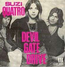 The front cover of Suzi Quatro's single Devil Gate Drive. Quatro is standing in the centre with her right hand on her hips and the other hand behind her. Standing behind her, on either side, are two members of her band. She is wearing a leather jacket, jeans, and many necklaces. The cover is in black and white with the words "Suzi Quatro" and "Devil Gate Drive" in hot pink.