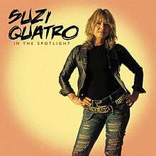 The front cover of Suzi Quatro's album In the Spotlight. Quatro is standing up with her hands on her hips. Behind her is yellow circle on an orange background, simulating the effect of a spotlight. She is wearing a black leather jacket (over a black top) and light blue jeans with horizontal holes ripped in them. She has two black leather belts - a plain one, plus one decorated with yellow studs and yellow chains.