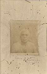 A faded sepia photograph of an old man with neck beads on a deteriorated paper page.