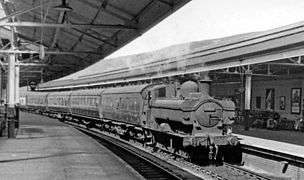 A pannier tank locomotive pulling five passenger coaches through a station